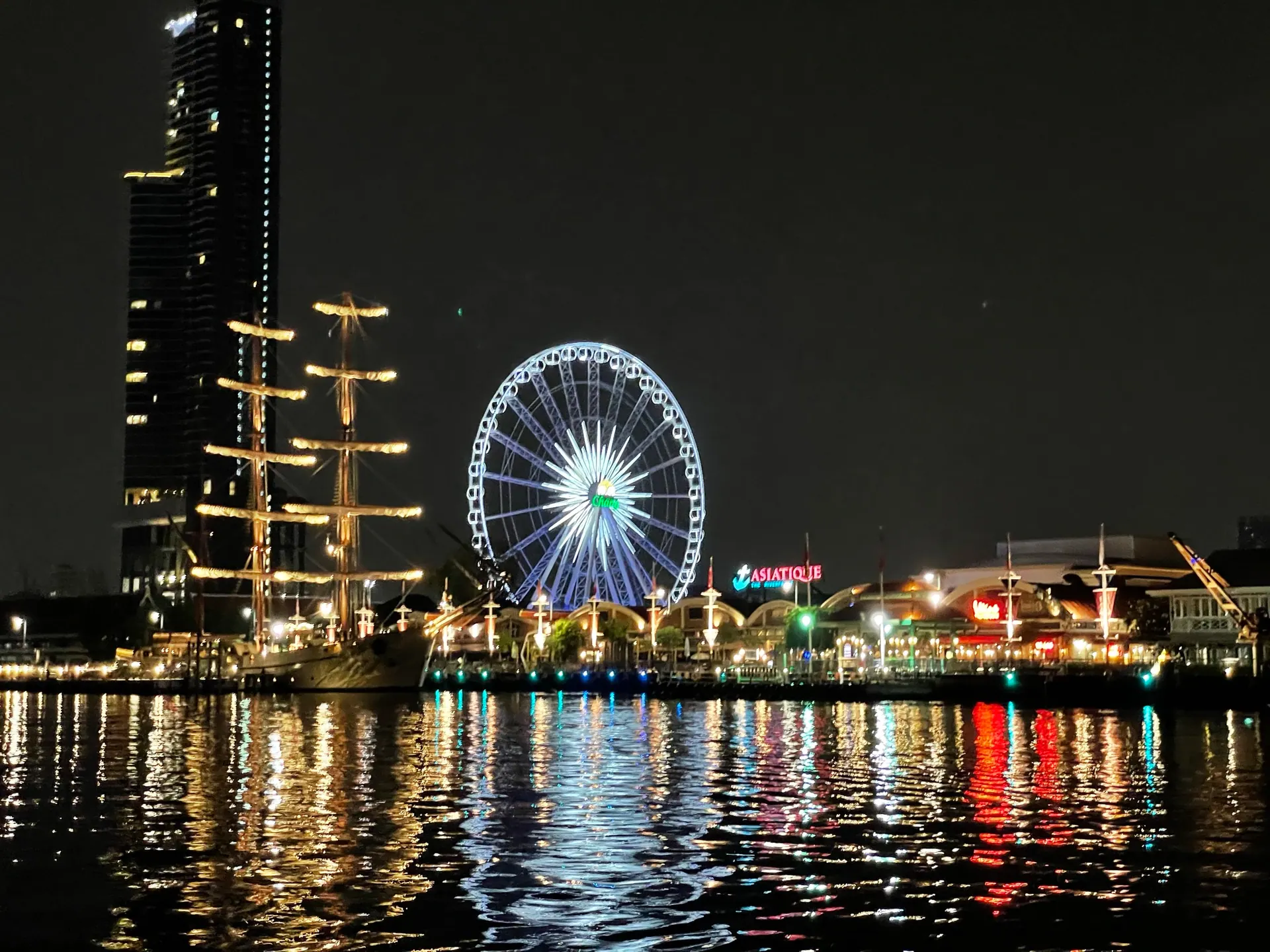 The Asiatique Ferris wheel and a tall ship lit up at night beside the river