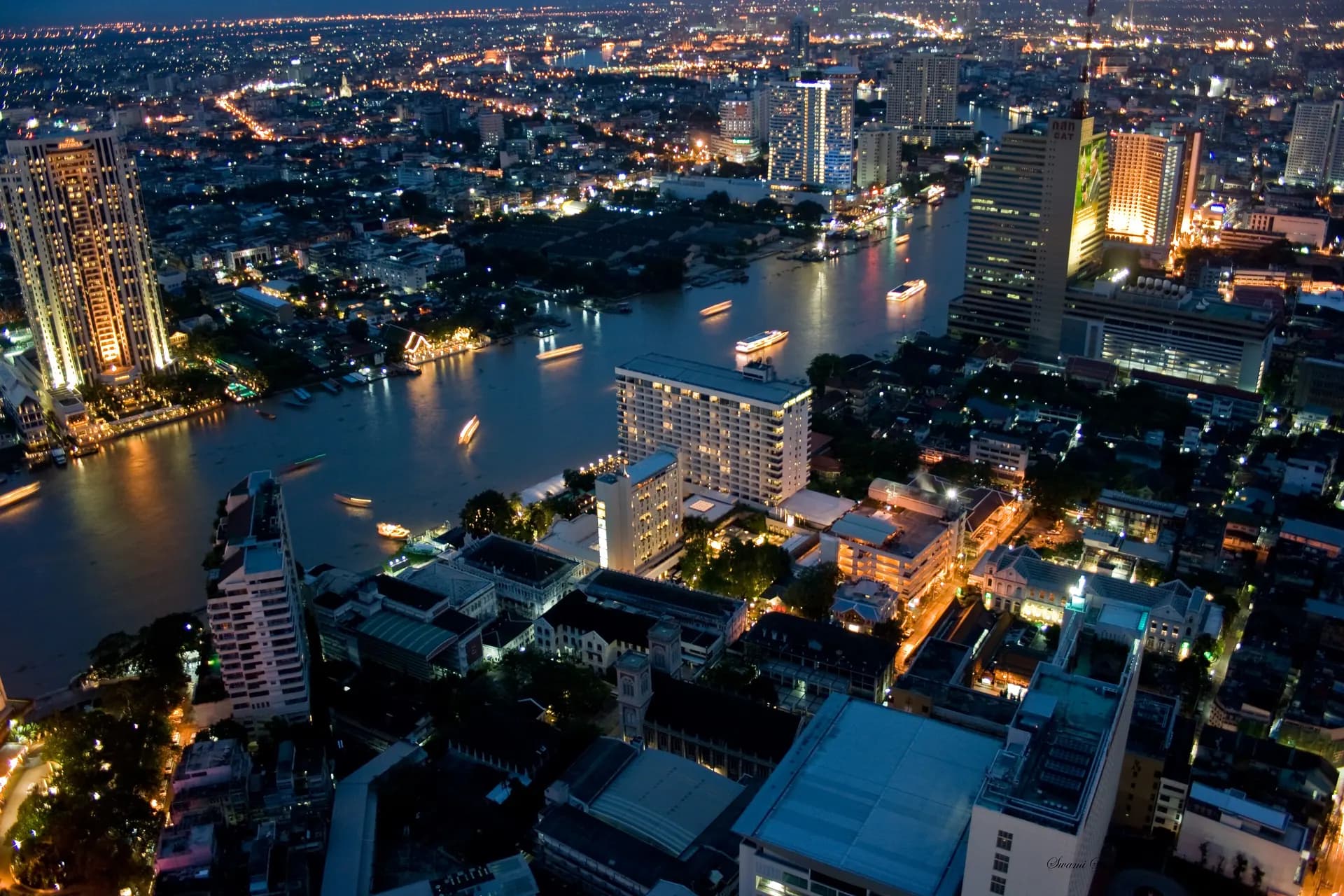 Bangkok at dusk seen from State Tower, with the Chao Phraya River and lit boats